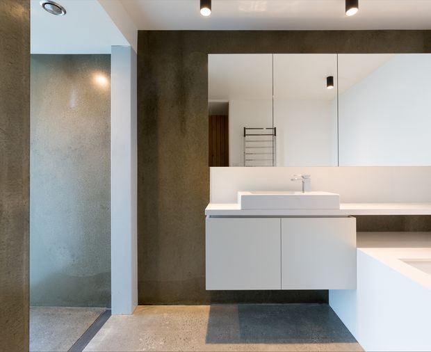 Contemporary minimalist bathroom with a floating white vanity, integrated rectangular basin, wall-mounted tapware, large frameless mirror, and a walk-in shower. Dark textured concrete-look wall surfaces contrast with white cabinetry and a pale benchtop. Recessed and black cylindrical ceiling lights provide focused illumination, while the floor combines large grey and light neutral tiles.