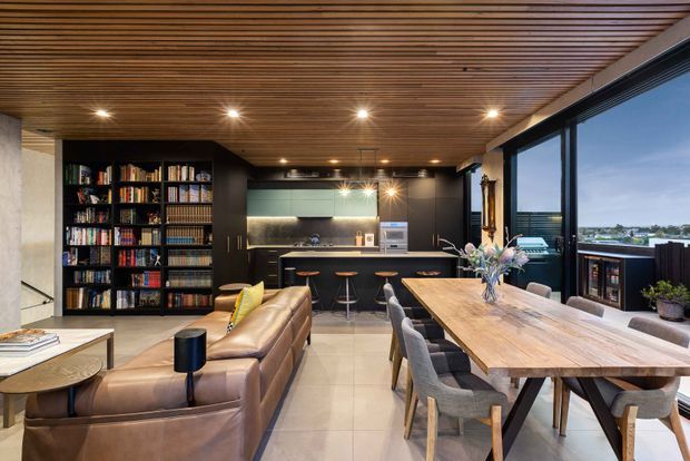 Open-plan contemporary living, dining and kitchen interior with a warm timber slatted ceiling, black built-in cabinetry and full-height glazing. A large timber dining table occupies the foreground, with a leather sectional sofa and extensive bookshelf joinery creating a connected living area. The kitchen has a dark finish with a pale green-grey splashback, and water is visible beyond the windows.
