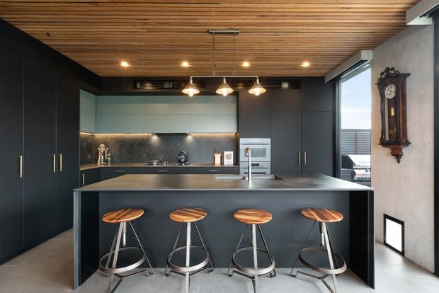 A contemporary kitchen with a large dark island and four stools, matte black cabinetry, integrated appliances, a reflective metallic splashback, and a warm timber slatted ceiling. Pendant lights hang over the island, while a tall window and traditional wall clock add contrast to the otherwise modern fit-out.