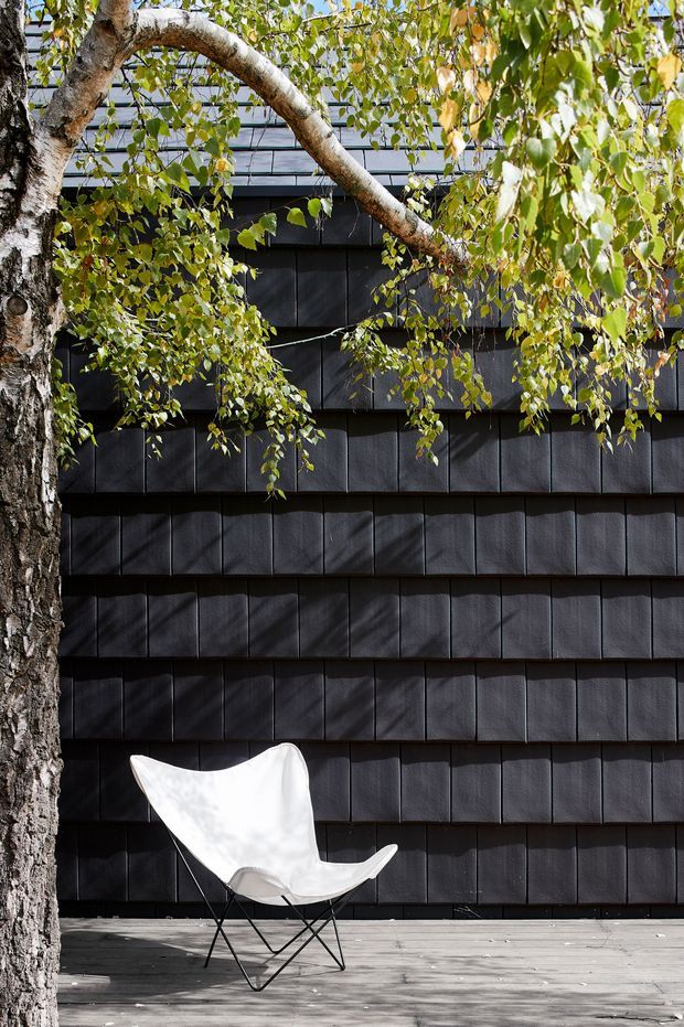 A close exterior view of a modern dark-clad building, partially screened by a leafy tree. The facade is covered in regularly arranged dark rectangular shingle-like panels, with horizontal bands creating a layered texture. A timber deck runs along the base, furnished with a white butterfly chair. Strong sunlight casts crisp tree shadows across the wall and deck.
