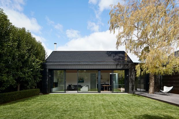 A single-storey dark-clad pavilion or house with a pitched tiled roof, broad floor-to-ceiling glazed doors, and a minimal modern interior visible through the glass. The building opens onto a neatly manicured lawn, with mature hedging and trees framing a timber deck and outdoor seating on the right.