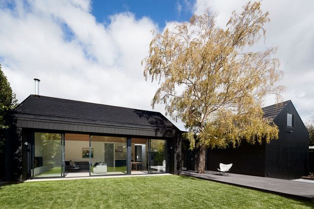 Exterior view of a modern single-storey home with dark charcoal cladding and a low-pitched gabled roof. Large floor-to-ceiling glazed doors open onto a broad lawn, while a connected dark-clad garage or secondary volume sits to the right beneath a steep gabled roof. A timber deck and mature tree frame the facade.