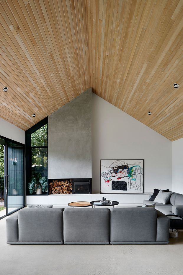 A contemporary living room with a large grey sectional sofa facing a minimalist fireplace and wood storage niche. The room features a dramatic pitched ceiling clad in narrow timber boards, smooth pale walls, polished concrete flooring, black-framed floor-to-ceiling glazing, and subdued modern furnishings. A large abstract artwork and indoor plants add visual interest.