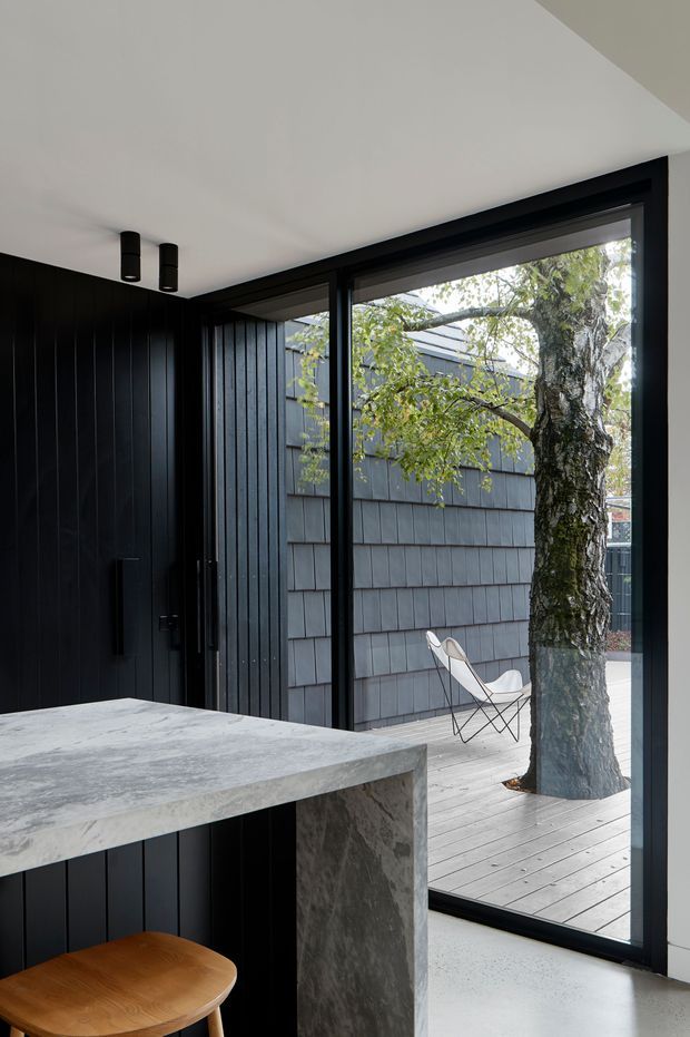 A refined modern kitchen interior viewed toward a full-height glazed opening and adjoining timber deck. The room features dark vertical wall panelling and cabinetry, a substantial grey stone island or peninsula, black-framed glazing, a white ceiling with compact black cylindrical downlights, and a pale polished or concrete-like floor. A mature tree, exterior cladding, and a small white outdoor chair are visible beyond the glass.