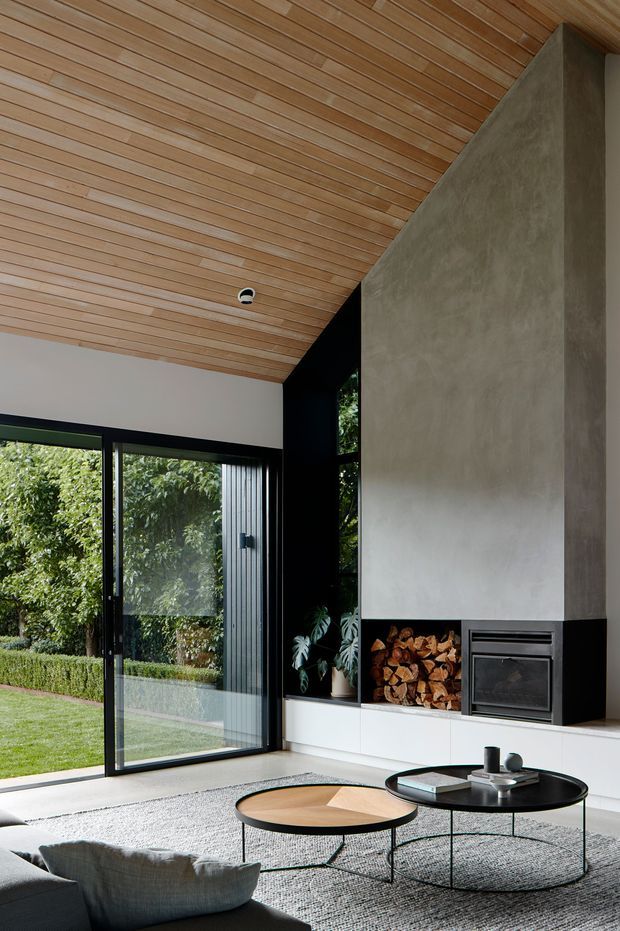 A contemporary living room with a dramatic full-height concrete or plaster fireplace wall, integrated wood storage, and black-framed floor-to-ceiling glazing opening to a garden. The room features a warm timber-lined sloping ceiling, pale built-in cabinetry, nesting round coffee tables, a large textured rug, and a dark sofa. The composition emphasizes the contrast between warm timber, pale surfaces, black detailing, and abundant garden views.