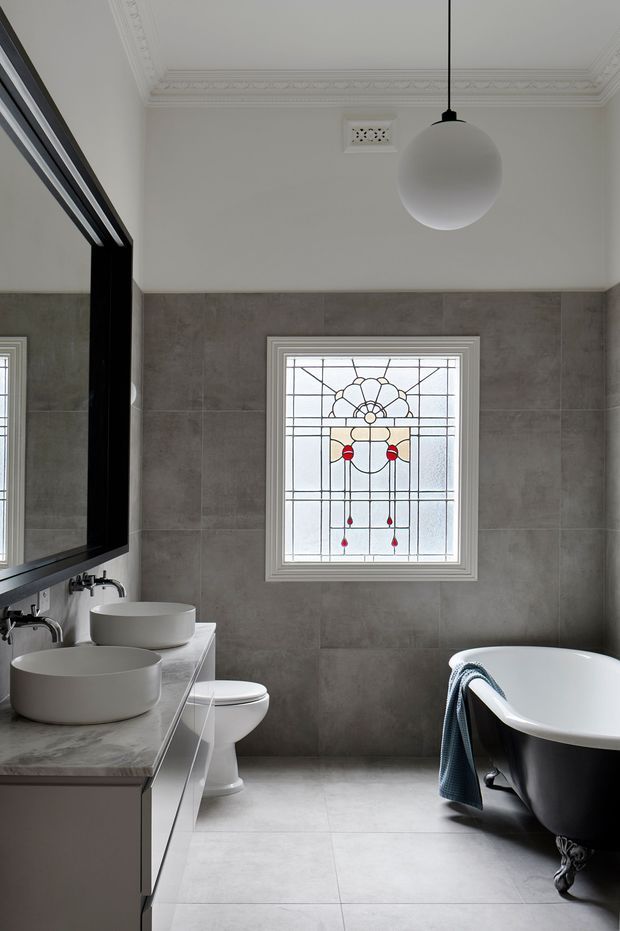 A refined bathroom with grey large-format tiled walls and floor, a pale stone vanity with two white vessel basins, a large black-framed mirror, toilet, and freestanding clawfoot bathtub. A decorative stained-glass window provides a colourful focal point, while ornate ceiling cornices and a globe pendant add traditional detailing. The palette is restrained in grey, white, and black with small red and yellow accents in the window.