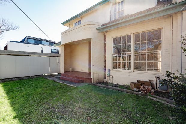 Exterior view of a traditional rendered and brick residential building, featuring a curved covered entry porch, large multi-pane timber-framed windows, a low-pitched roof with exposed weathering, and a small grassed yard. A modern neighboring structure is visible beyond the boundary fence.