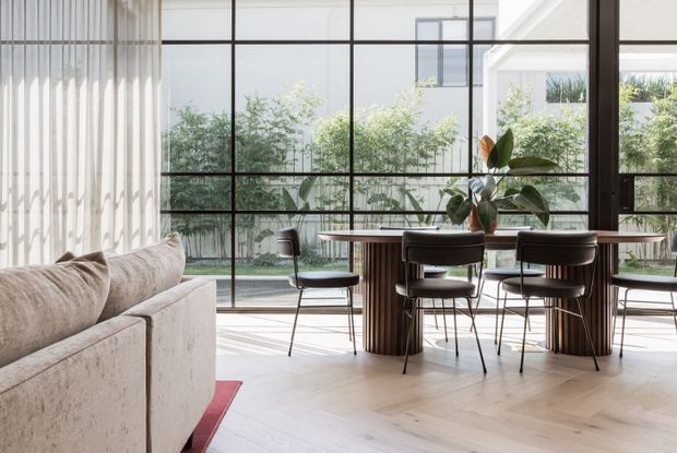 Bright contemporary dining and living area with a large black-framed glass wall overlooking a landscaped courtyard. A round dining table with dark upholstered chairs sits on pale herringbone timber flooring, while a neutral textured sofa occupies the foreground. Sheer curtains, indoor plants and strong natural light create an airy, refined interior.