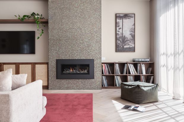 A bright, contemporary living room with a built-in fireplace clad in small patterned mosaic tiles, a wall-mounted television, timber shelving filled with records, and a low timber media cabinet. A pale upholstered sofa, deep red area rug, leather beanbag, indoor plant, and framed artwork furnish the room. Full-height sheer curtains provide abundant natural light, with pale timber flooring throughout.