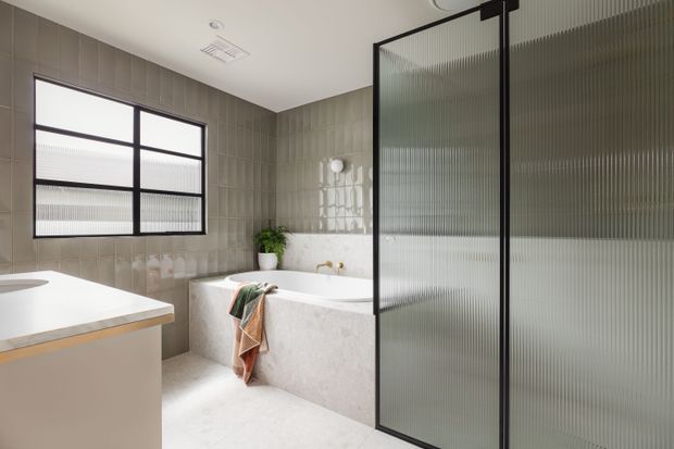 A bright contemporary bathroom featuring a freestanding white bathtub, a white vanity with a pale stone benchtop, large grey-beige rectangular wall tiles, pale flooring, and a black-framed ribbed glass shower screen. A black-framed window provides natural light, while a small plant and towels add modest warmth.