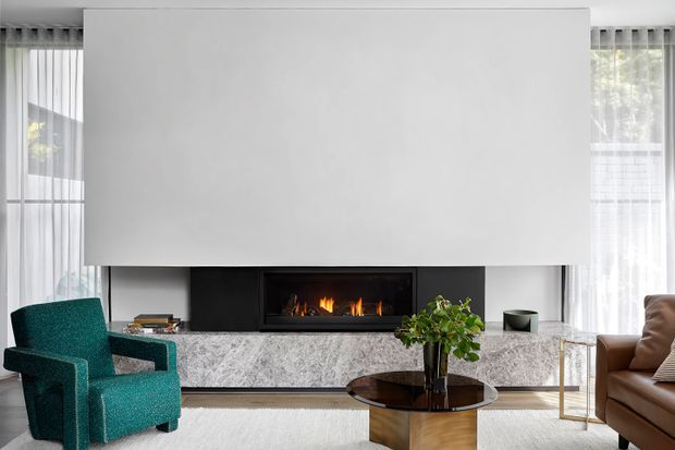 Contemporary living room with a large white feature wall and integrated black fireplace, a grey marble fireplace surround and hearth, sheer floor-to-ceiling curtains, teal accent armchair, brown leather sofa, and a round dark coffee table. The room has a refined, warm, high-contrast interior with abundant natural light.