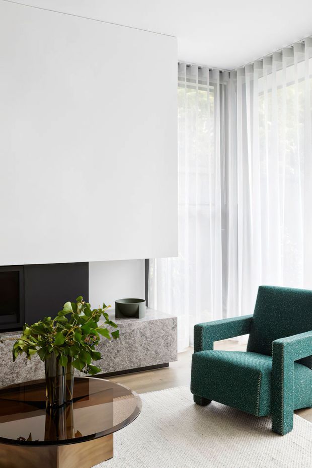 Bright contemporary living room with a large white built-in media wall and suspended-looking white volume, dark recessed fireplace or television niche, grey stone-effect low cabinetry, floor-to-ceiling sheer curtains, a teal upholstered armchair, glass coffee table, rug, and indoor greenery. The room has a clean, refined, minimalist feel with abundant filtered daylight.