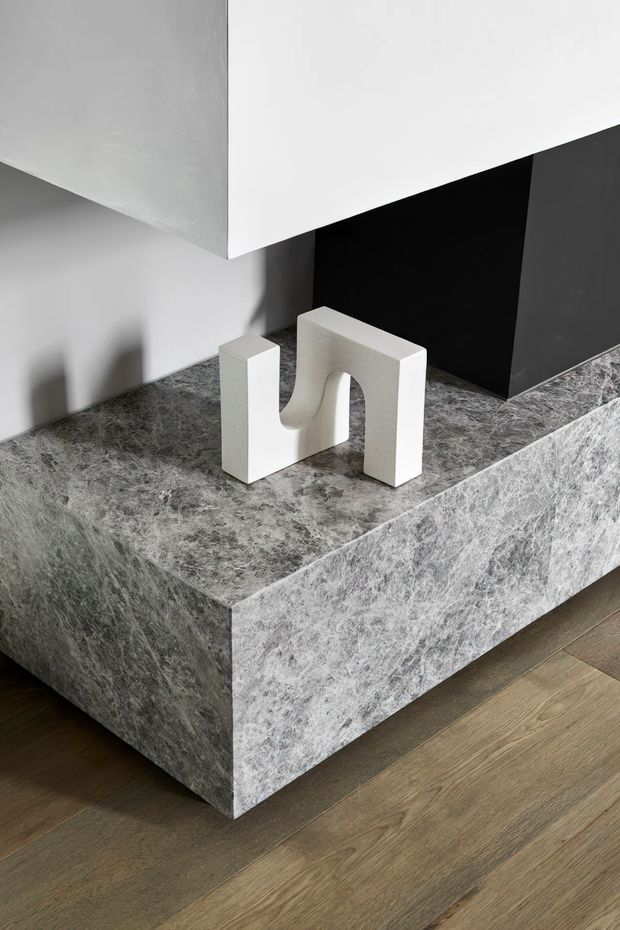Minimalist interior detail featuring a substantial grey veined stone-clad plinth or low built-in console, set against a black and white geometric wall composition. A small white sculptural object sits on top, with warm timber flooring below.
