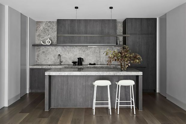 A compact contemporary kitchen with dark timber-look cabinetry, a grey stone-textured splashback, a matching island with two white bar stools, dark timber flooring, and minimalist pendant lighting. The space has a clean, refined monochromatic palette with warm wood tones.