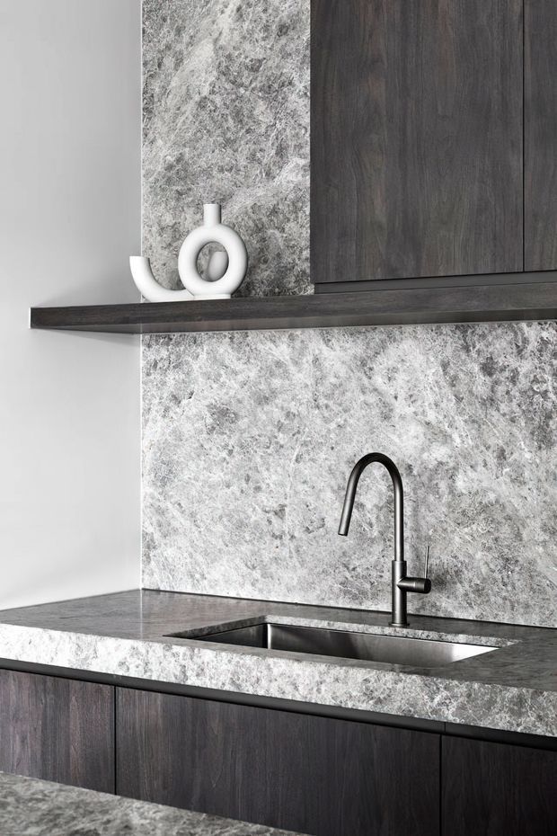 A close-up of a contemporary kitchen featuring dark timber cabinetry, a dramatic grey-and-white natural stone splashback and benchtop, an undermount stainless-steel sink, and a matte black gooseneck tap. A slim dark timber floating shelf with minimalist white ceramics is mounted above the benchtop.