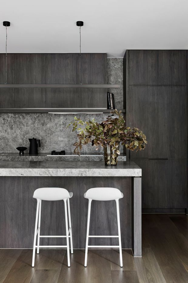 A refined contemporary kitchen with dark timber cabinetry, a mottled grey stone backsplash and island benchtop, integrated shelving, black pendant lights, and two white bar stools. A tall dark cabinet and timber flooring create a cohesive, moody palette, softened by a leafy arrangement on the island.