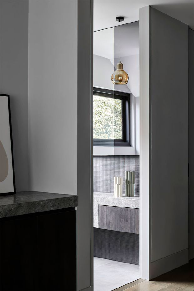 A narrow view through a partially open doorway into a refined bathroom. The room features a dark-framed window, small grey mosaic wall tiles, a pale stone vanity benchtop with integrated cabinetry, and a warm amber pendant light. Dark cabinetry and stone surfaces in the foreground create a sophisticated, monochromatic transition between spaces.