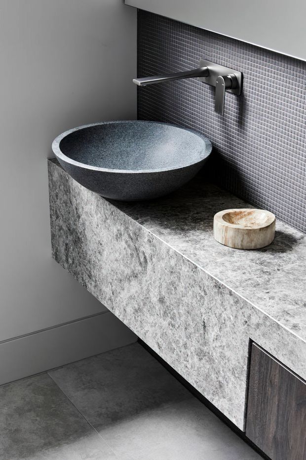Close-up of a refined contemporary bathroom vanity with a round dark stone vessel basin, wall-mounted brushed metal tapware, a large grey stone benchtop and matching waterfall-style vanity front, dark timber cabinetry, grey floor tiles, and a small dark mosaic-tile splashback. The composition is monochromatic and highly material-focused.