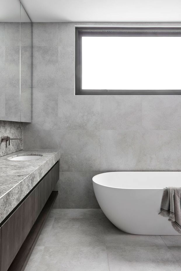Contemporary minimalist bathroom featuring large-format grey stone-look wall and floor tiles, a floating dark timber vanity with a pale stone benchtop and integrated basin, wall-mounted tapware, a frameless mirrored cabinet, and a freestanding white bathtub. A narrow horizontal window provides natural light, while a neutral grey palette creates a calm, refined atmosphere.