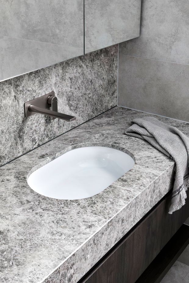 Close-up of a refined contemporary bathroom vanity featuring a pale grey veined stone benchtop with an integrated oval white basin, a wall-mounted brushed dark-metal tap, matching grey stone-look wall surfaces, and dark timber cabinetry below. A textured grey towel rests on the benchtop.