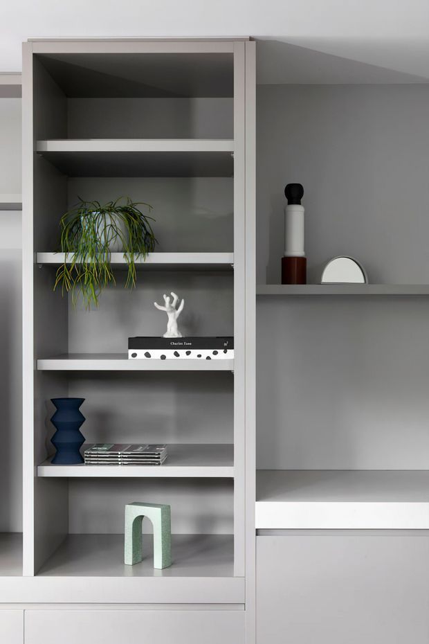 Close-up interior view of a built-in grey shelving and cabinetry unit. Open shelves display a trailing plant, books, sculptural objects, and decorative vessels, while adjacent recessed shelves provide additional display space. The joinery has clean rectangular lines, a muted grey palette, and a minimalist contemporary appearance.