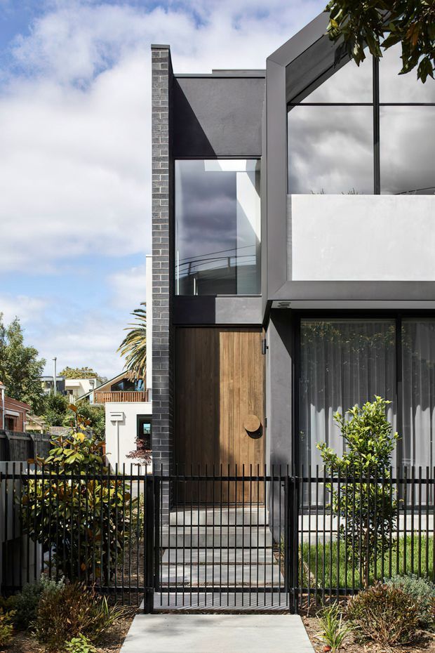 Street-facing view of a narrow contemporary multi-storey residence with a strong geometric facade. The home combines dark brickwork, charcoal rendered surfaces, warm vertical timber cladding, expansive black-framed glazing, and a projecting upper-level balcony or enclosed terrace. A black metal front fence, concrete entry path, and compact landscaped garden frame the entrance. Neighbouring suburban homes and vegetation are visible in the background.