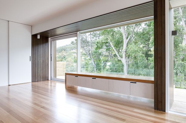A bright, minimalist interior with expansive floor-to-ceiling glazing overlooking leafy surroundings. The room features pale timber flooring, white walls, dark vertical timber framing and panelling around the windows, and a long built-in low cabinet beneath the glazing. A timber deck or exterior terrace is visible beyond the doors.