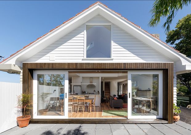 Bright front exterior view of a renovated or extended gable-roofed home. White horizontal weatherboard cladding and white trim surround a large glazed opening framed in dark timber, with sliding doors opening onto a paved terrace. The interior visibly includes a kitchen and living area, while the exterior foreground features pots and paving.