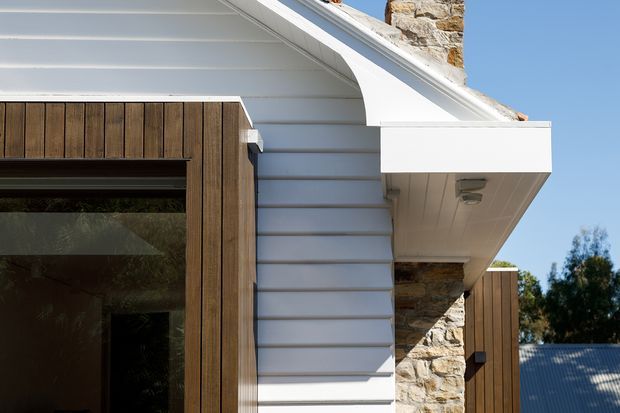 Close-up exterior architectural detail showing a white gabled roof edge and eaves, horizontal white weatherboard cladding, natural stone masonry, and a large dark-framed glazed opening bordered by vertical timber cladding. The composition highlights the junction of contemporary timber and traditional stone and weatherboard elements under clear daylight.