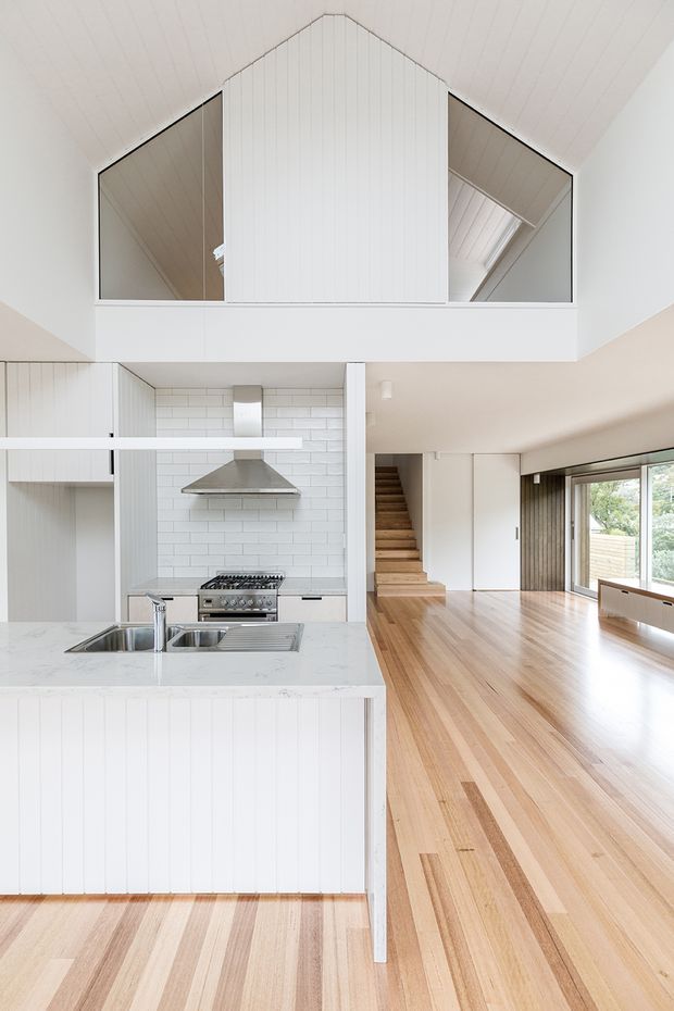 Bright open-plan interior centered on a white kitchen with a large marble-look island, stainless-steel sink and rangehood, white tiled splashback, and vertical white wall panelling. The kitchen opens directly into a spacious timber-floored living area with large glazing, a timber staircase, and a dramatic double-height pitched ceiling. A mezzanine-like upper opening with mirrored panels and white cladding overlooks the room.