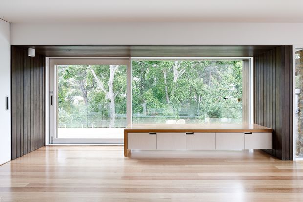 A bright, minimalist interior with a large horizontal picture window overlooking greenery. The room features pale timber flooring, dark vertical timber wall panelling around the window, white walls and ceiling, and a long built-in low cabinet with a warm timber top beneath the glazing. The space appears open and uncluttered, with strong horizontal lines and abundant natural light.