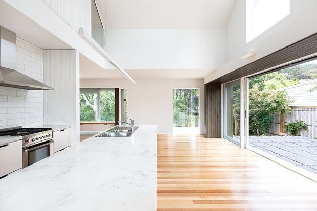 Bright open-plan kitchen and living area with a long white stone island, stainless-steel appliances, white subway-tile splashback, pale timber flooring, high white ceilings, and extensive sliding glass doors opening to a paved outdoor terrace. The space has generous natural light and a strong indoor-outdoor connection.