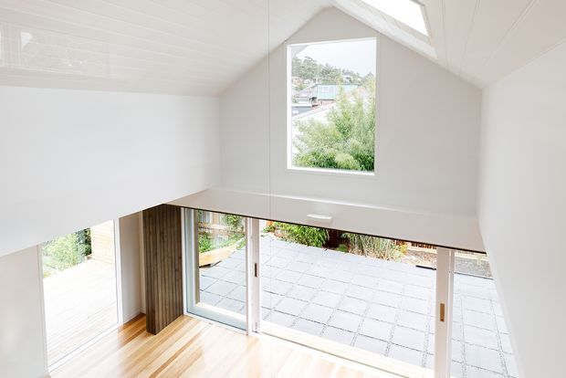 Bright compact interior with a high pitched, white-lined ceiling and clerestory-style upper window. Large glazed sliding doors open onto a paved courtyard or patio, while warm timber flooring and vertical timber wall detailing add natural contrast to the predominantly white space. The elevated viewpoint and open volume suggest a loft-like living area.