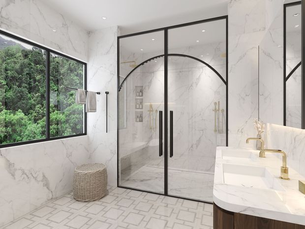 A bright, luxurious bathroom clad in white marble-look surfaces, featuring a glass shower enclosure with distinctive black metal framing and arched detailing. The room includes a white double vanity with a curved marble-look benchtop, brushed brass fixtures, wall-mounted towel rails, and a woven laundry basket. Large windows overlook dense greenery, while the patterned light floor adds visual interest.