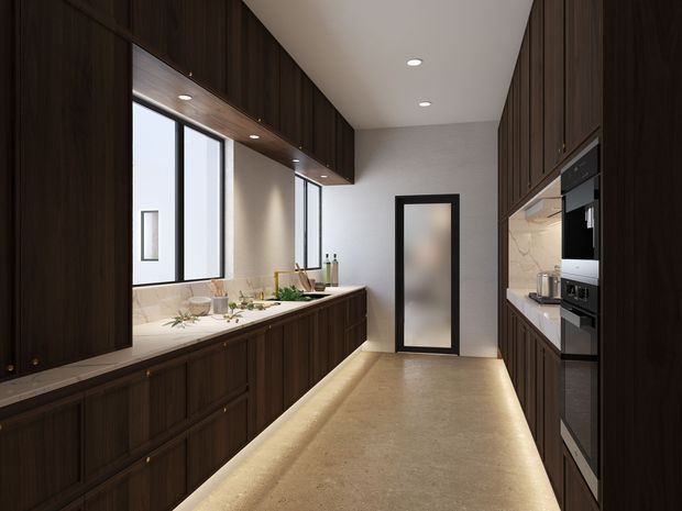 A narrow galley kitchen with dark timber cabinetry on both sides, integrated black appliances, a pale stone benchtop and backsplash, tall windows, recessed ceiling lighting, and warm under-cabinet illumination. A frosted-glass door forms the rear wall, while the central aisle is finished in a light stone or concrete-like surface.