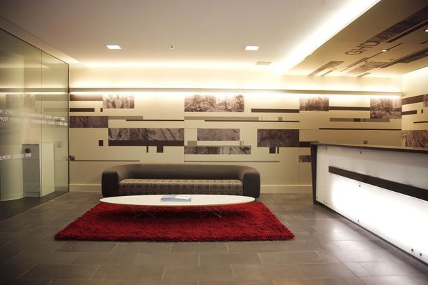 A contemporary commercial reception lobby with a long illuminated reception desk, dark upholstered sofa, low oval coffee table, and red area rug. The space features grey tiled flooring, a cream-toned feature wall with layered horizontal graphic panels and monochrome photographic artwork, mirrored glazing on the left, and recessed ceiling lighting with concealed perimeter illumination.