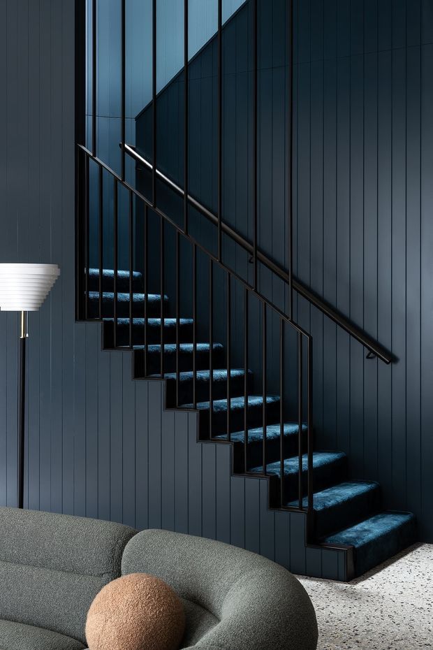 A moody contemporary interior featuring a floating staircase with dark timber or painted vertical wall panelling, slim black metal balustrades, and deep blue carpeted treads. A curved grey upholstered sofa with a warm tan cushion occupies the foreground, while a sculptural floor lamp is partially visible at left. The floor appears to be light terrazzo or speckled concrete.