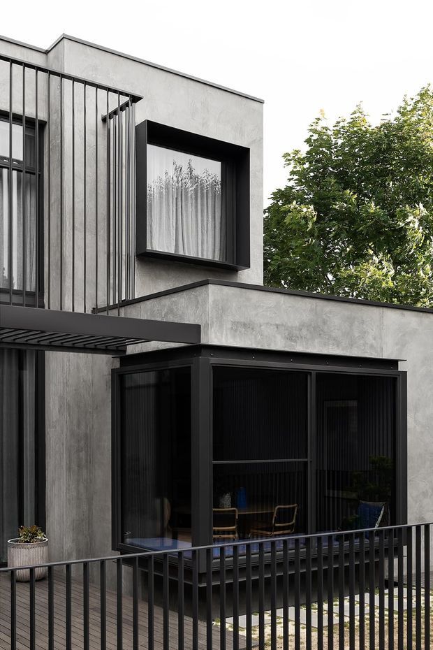 Exterior view of a contemporary multi-level residence with smooth grey rendered walls, strong black metal framing, vertical metal screening, and large dark-framed glazed openings. A covered balcony or terrace with black railings occupies the foreground, while trees are visible behind the building.