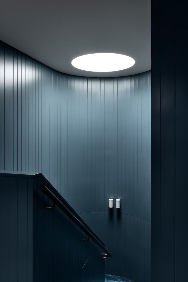 A dramatic, dark stairway interior with deep blue-grey vertically grooved wall panelling, a curved ceiling, and a large circular recessed ceiling light. A black angular stair balustrade descends through the foreground, with small wall-mounted cylindrical lights providing subtle illumination.