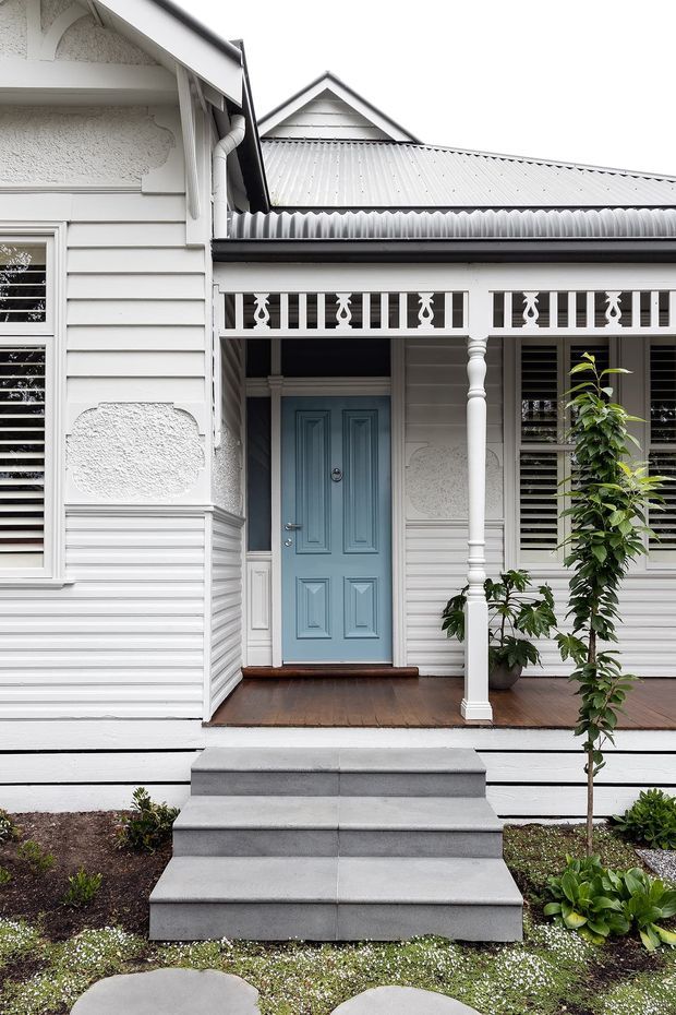 Front entry of a restored or traditionally styled weatherboard house, featuring white horizontal cladding, decorative plaster panels, a pale blue paneled front door, timber verandah decking, ornate fretwork balustrade, turned timber posts, louvred windows, and broad concrete steps. Low garden planting borders the approach.