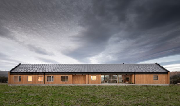 Long, single-storey timber-clad building set in an open grassy landscape, with a simple symmetrical pitched metal roof, regularly spaced windows, and a glazed central entrance or living area. Dramatic overcast sky and distant wooded hills form the background.