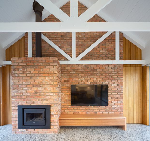 Interior view of a contemporary living room centred on an exposed red-brick fireplace and matching brick feature wall. A black built-in wood-burning fire sits within a projecting brick hearth, with a wall-mounted television above a low timber media cabinet. White-painted post-and-beam trusses form a prominent structural feature beneath a pitched ceiling, while vertical timber wall panelling frames both sides. The floor is a light terrazzo-like or aggregate finish.