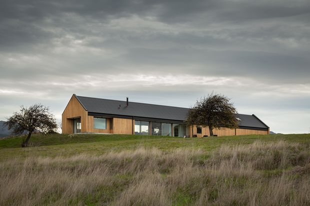 A low, elongated contemporary rural house set in an open grassy landscape beneath an overcast sky. The home features dark pitched gable roofs, warm vertical timber-clad walls, extensive glazed openings, and a simple single-storey form. Sparse trees and rolling terrain surround the building.