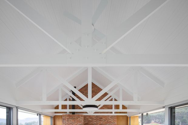 Interior view of a bright, open room with a high pitched timber-lined ceiling and exposed white-painted roof trusses. A central white ceiling fan hangs above the space, while large glazed openings on both sides provide expansive views. The far wall features exposed red brick with warm timber or wood-toned vertical elements, creating a light yet rustic architectural character.