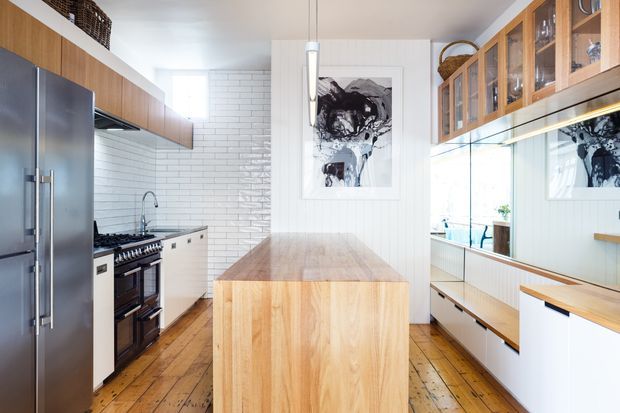Bright galley-style kitchen with a central timber island, stainless-steel double refrigerator, freestanding dark range cooker, white subway-tile splashback, timber flooring, white lower cabinetry, and extensive glazed timber display cabinetry. A mirrored wall visually expands the room, while abstract artwork and woven storage add decorative detail.