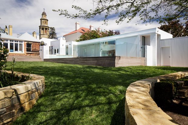 Exterior view of a contemporary white-clad residential extension set within a landscaped lawn and bordered by low sandstone retaining walls. The building has extensive floor-to-ceiling glazing, a long low horizontal form, a shallow pitched or low-profile roof, and a dark timber-clad base. A more traditional brick and rendered heritage building with a prominent tower is visible behind it, creating a clear contrast between old and new architecture.