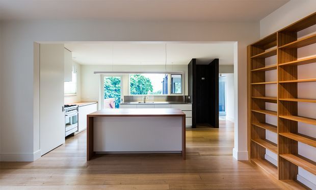 A bright, open-plan kitchen viewed from an adjoining living or dining area. The kitchen features a central island with a dark timber base and pale benchtop, white cabinetry, integrated appliances, timber flooring, a black full-height pantry or joinery unit, and a large built-in timber shelving wall in the foreground. Natural light enters through the rear windows, creating a clean, warm contemporary interior.