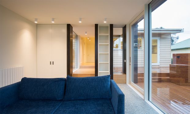 A contemporary living room with a deep blue sofa, pale built-in cabinetry, light carpet, recessed ceiling lights, and extensive floor-to-ceiling glazing. The glazing opens views to a timber deck and a neighbouring low-rise structure, while a central built-in shelving and partition element leads toward a brightly lit hallway.
