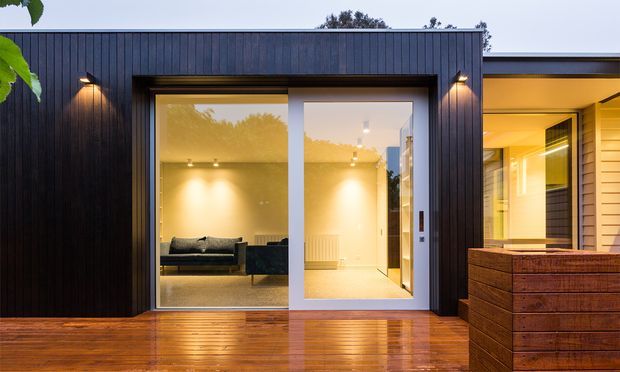 A contemporary residential rear facade viewed from a timber deck at dusk. The single-storey extension is clad in dark vertical timber boards and has a large glazed sliding door opening into a brightly lit living room. Warm exterior wall lights illuminate the facade, while a timber privacy screen or planter structure sits at the right. A lighter weatherboard-clad section is visible beyond the extension.