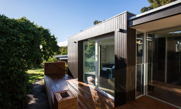 Exterior view of a contemporary single-storey residence with dark vertical timber cladding, extensive glazing, and a warm timber deck. The deck includes built-in timber seating or planter-like boxes, while dense hedging and mature greenery frame the private outdoor space. Strong sunlight creates a warm, inviting connection between the interior and the landscaped exterior.